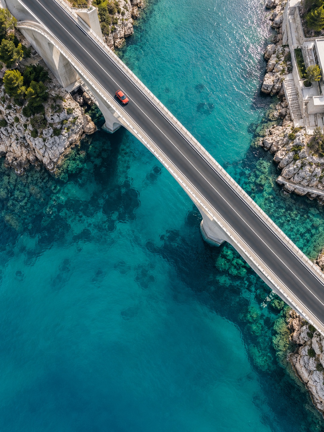 A top-down aerial view of a modern bridge and red car over turquoise coastal water.