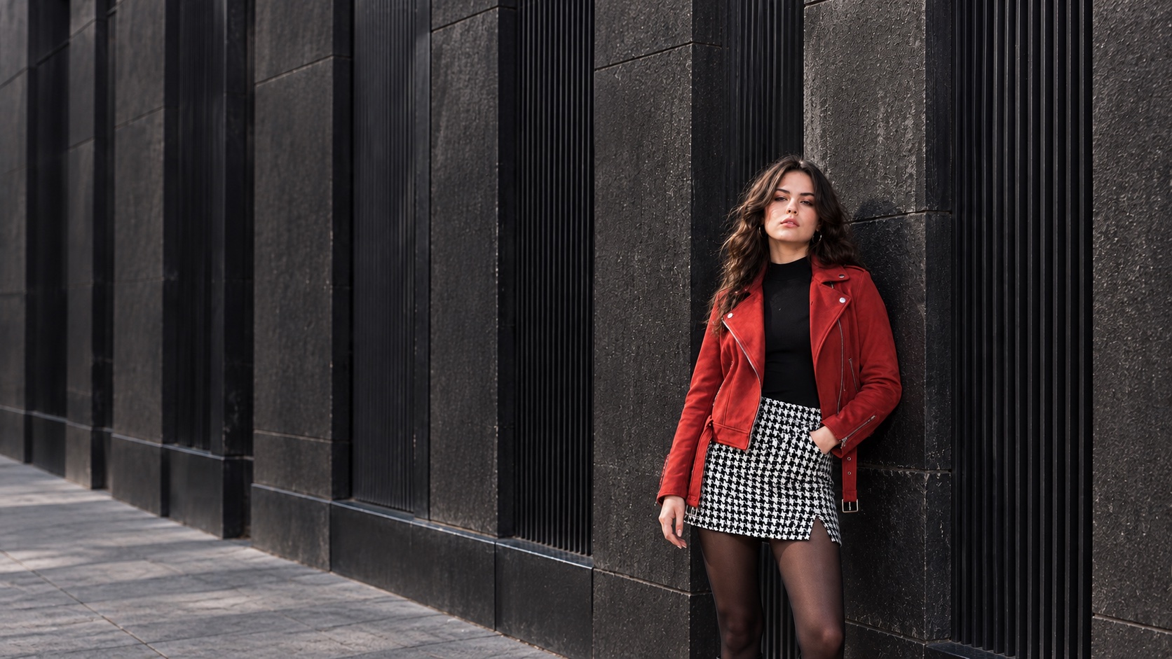 A woman in a red suede jacket beside a charcoal urban facade, in a wide editorial composition.